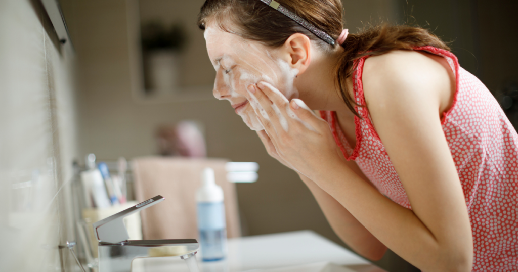 Teen girl gently washing her face with a foaming cleanser at a bathroom sink.
